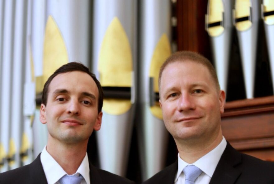 Headshots of two men smiling with closed mouths in front of a cathedral organ. 