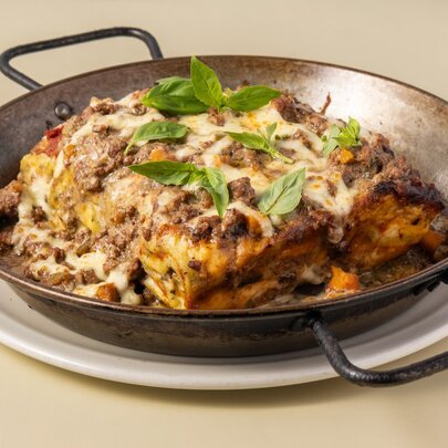A large serve of lasagne, topped with melted cheese and basil leaves, in a cast iron pan, resting on a tabletop.