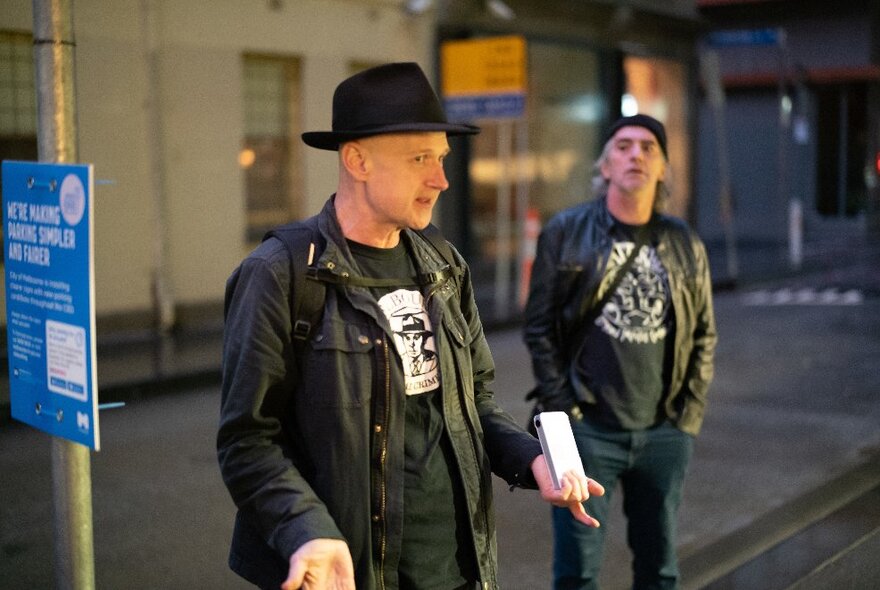 A man wearing a black hat and talking, leading a walking tour through the city of Melbourne, with a tour participant listening.