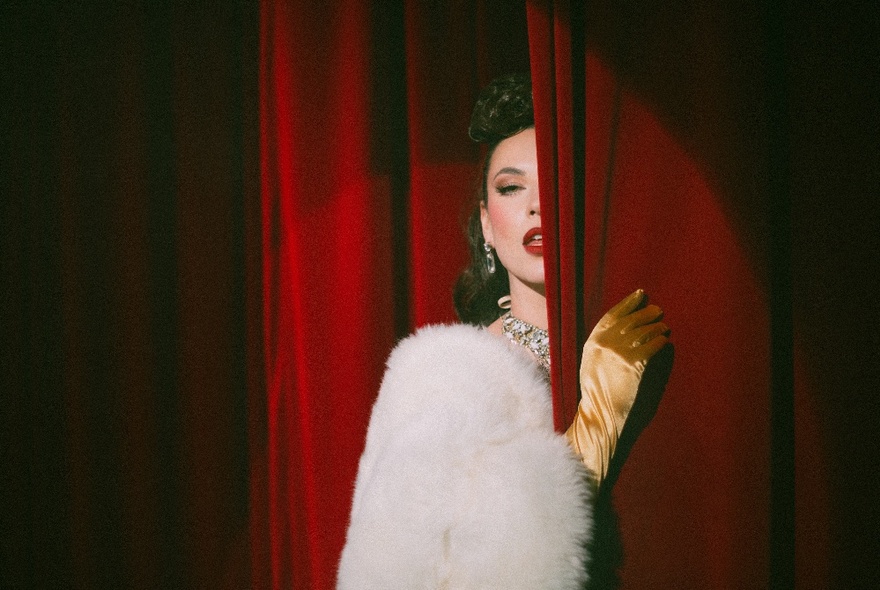 A glamorous woman wearing a white fur jacket and gold satin gloves, hiding behind a red velvet curtain.