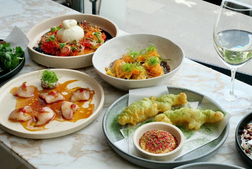 Four plates of food on a restaurant table at Taxi Kitchen, with a glass of white wine.