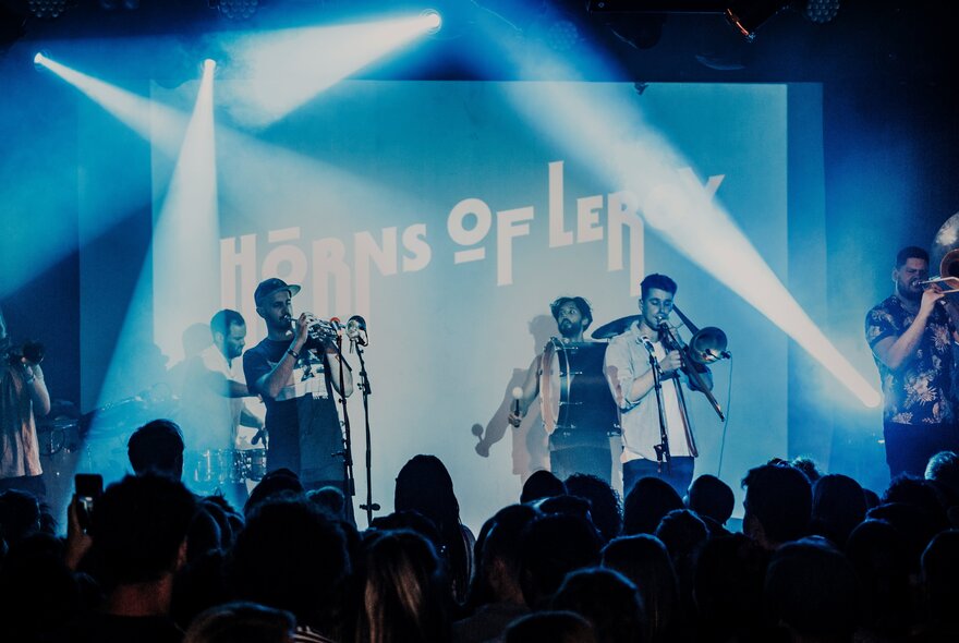 A horn band on a blue-lit stage with stage lighting, seen from behind a darkened audience.