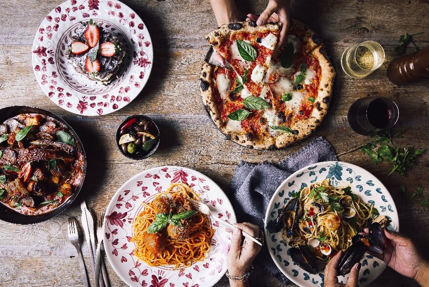 Dishes of pasta and pizza on a wooden table with drinks.