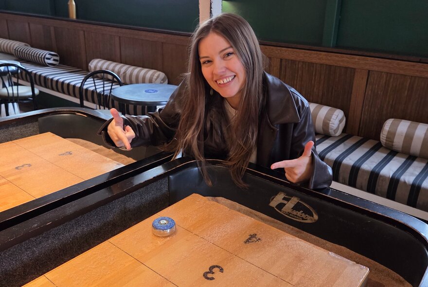 Smiling woman seated at a table in a venue, pointing at a buzzer.