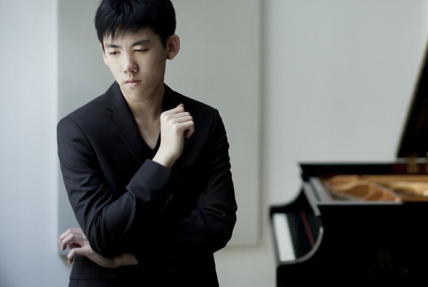 Pianist Haochen Zhang wearing a black suit jacket and looking reflective, a grand piano behind him.