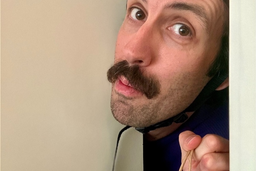 A man with a moustache peeking around a door with a helmet strap under his chin. 