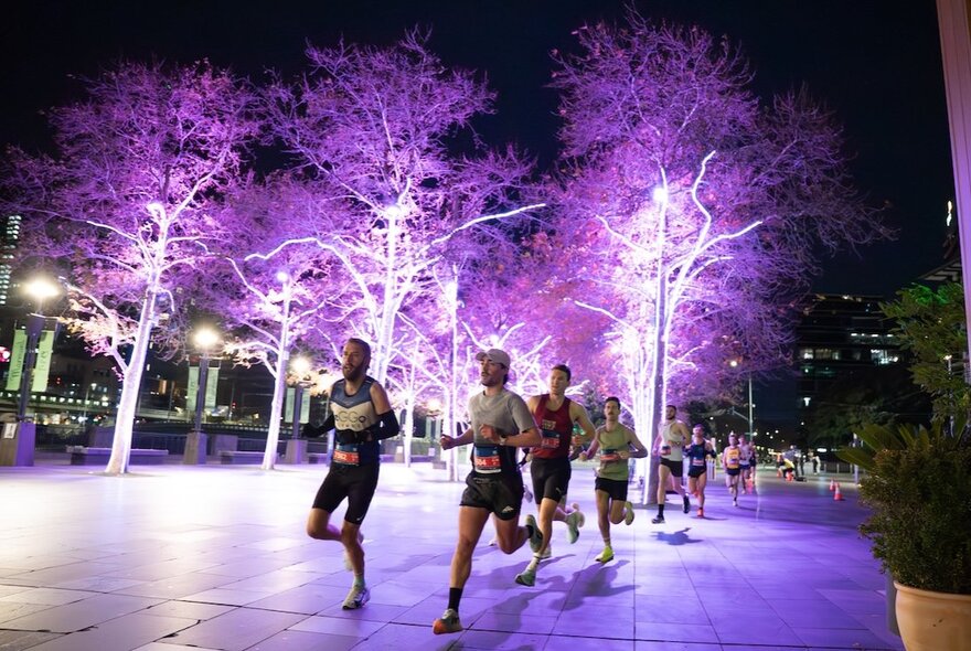 People running at night, on a paved path past trees lit up with purple lights, as part of an organised event.