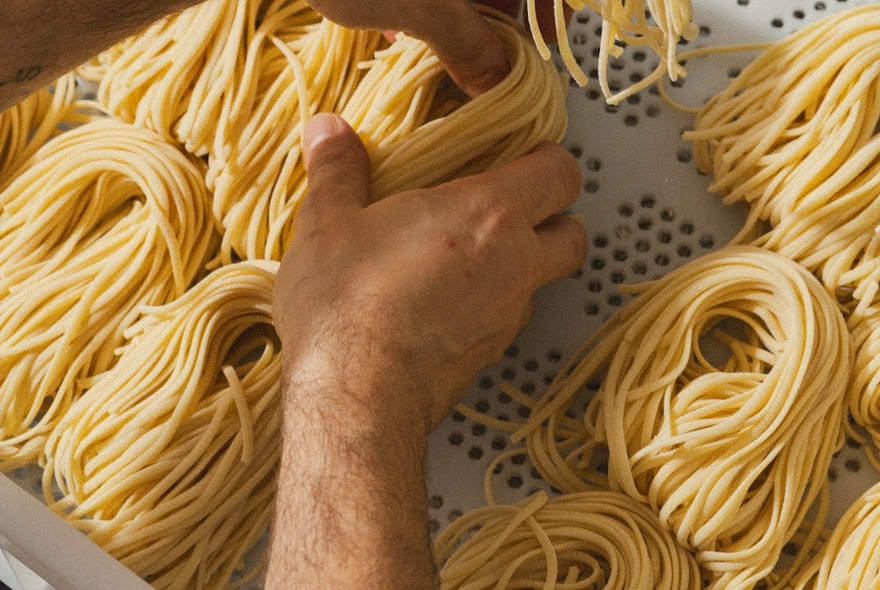 Neat piles of hand-made spaghetti on a tray; a person's hand placing a pile down.