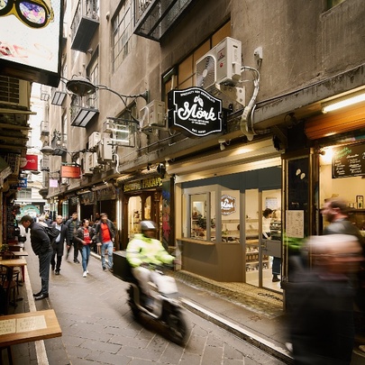 Centre Place laneway showing the brightly lit shop exterior of Mork Chocolates, with hanging signage, and pedestrians walking past.