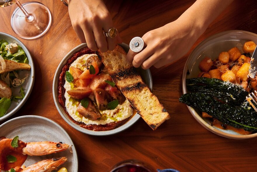 Overhead view of a table in a restaurant with a hand breaking a piece of bread over a plate of food, with other dishes nearby.