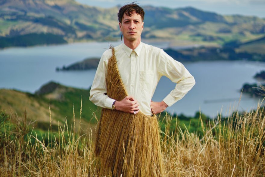 Singer Marlon Williams standing in the landscape with a lake and mountains in the background, wearing a white shirt with a grass skirt draped over one shoulder.