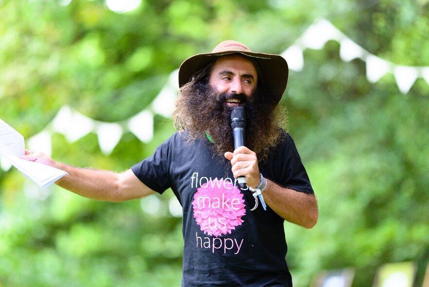 A man with a very bushy beard and wide-brimmed hat, presenting into a microphone. 