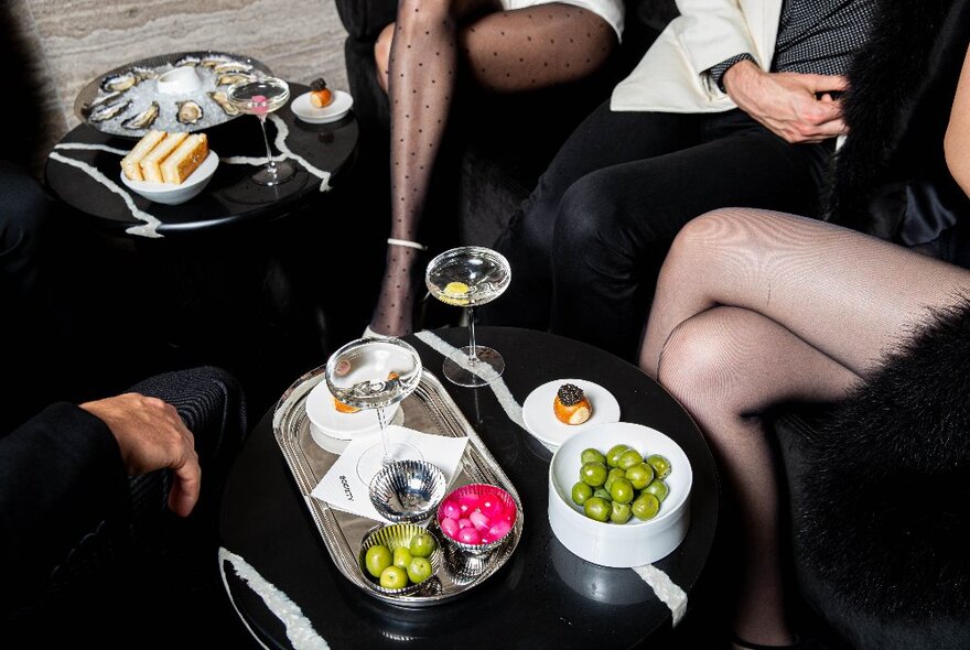 The legs of three people seated around a small coffee table that holds a platter of high-end snacks and olives and three glasses of martini.