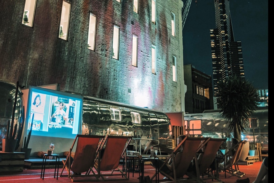 Row of deck chairs facing a small outdoor cinema screen, with a vintage 1970s silver airstream trailer positioned against the side of a building.