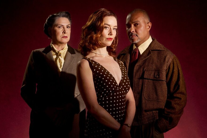 Three actors in period dress, an older woman in a tweed suit, a man in a brown coat and a young woman with a revealing polka-dot dress.