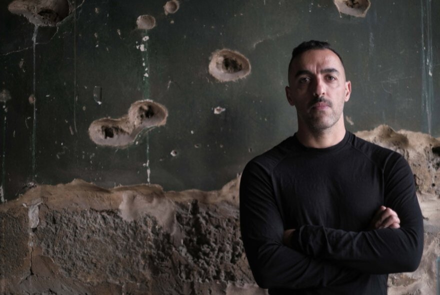 Antoun Issa, a man standing with his arms crossed in front of a dark, war-torn wall.

