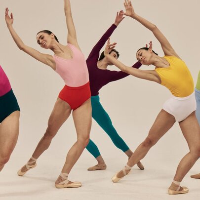 Five ballet dancers in a group pose, leaning gracefully to their right with arms elegantly curved overhead, all dressed in vibrant, contrasting dancewear.