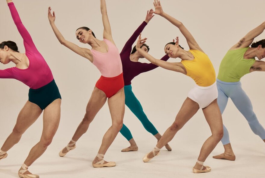 Five ballet dancers in a group pose, leaning gracefully to their right with arms elegantly curved overhead, all dressed in vibrant, contrasting dancewear.