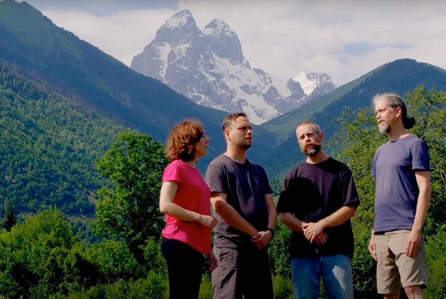 Four people standing outdoors, high snow capped mountains and green hills behind them.
