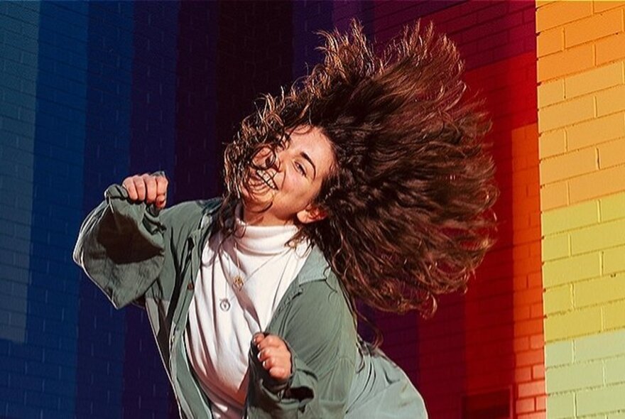 A woman dancing in front of a rainbow-coloured brick wall, her long brown hair fanning out around her.