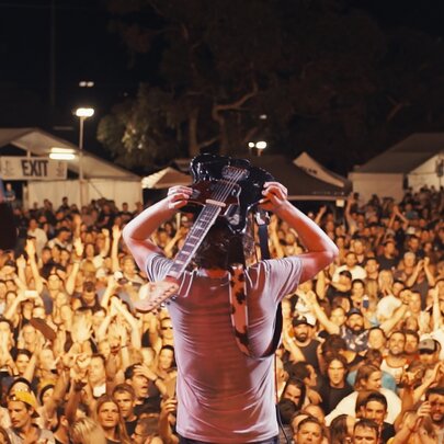 A still from the documentary film Jebediah: Are We OK? features lead singer Kevin Mitchell balancing a guitar on his head, standing on a stage in front of a large audience at night.