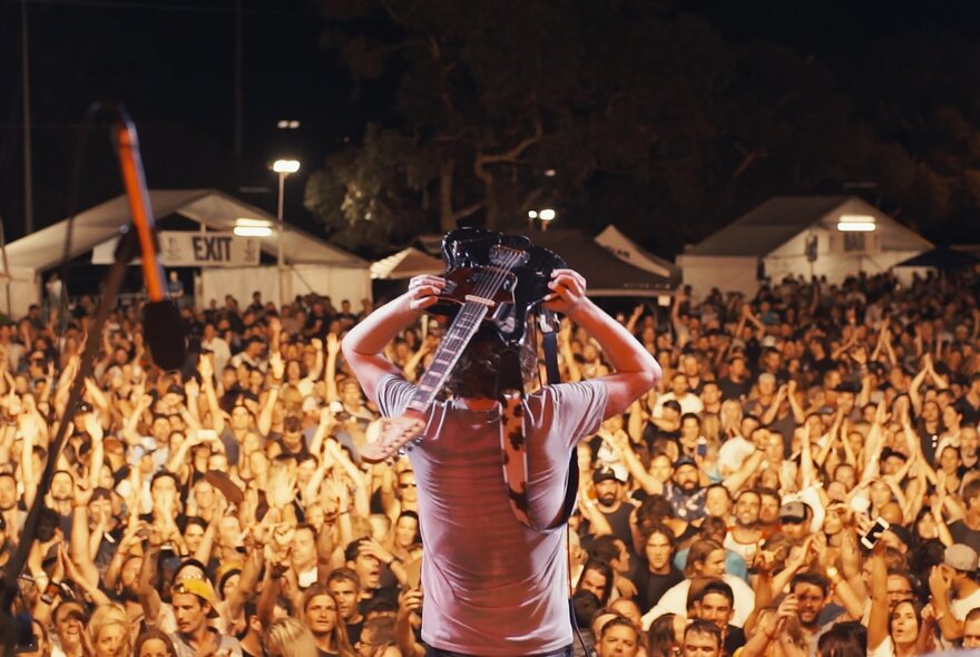 A still from the documentary film Jebediah: Are We OK? features lead singer Kevin Mitchell balancing a guitar on his head, standing on a stage in front of a large audience at night.