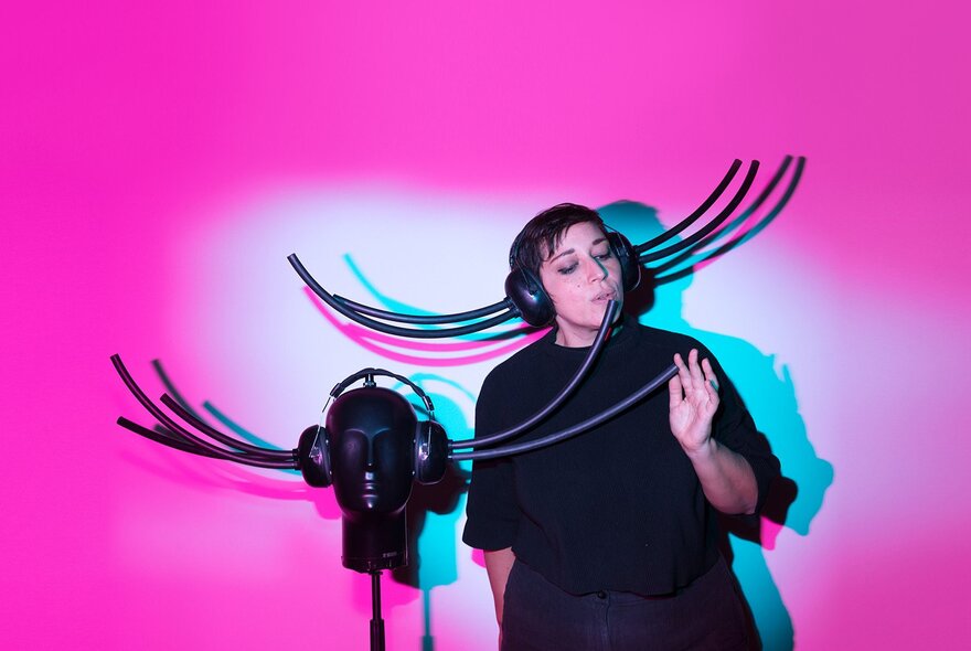 A person wearing headphones with plastic tubing attached directly to the earpieces, making sounds with her mouth into plastic tubing attached to another set of headphones on a dummy's head.
