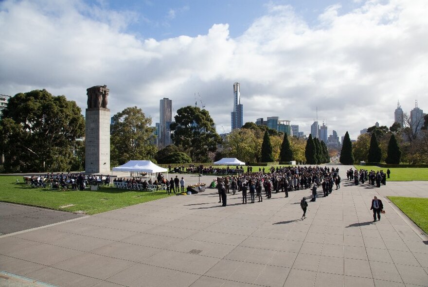 People in army uniform standing at a public ceremony in a park area with the city skyline in the distance. 