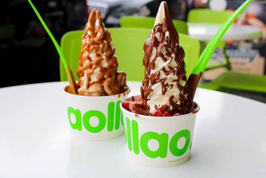 Green spoons being dipped into two small white cups with lime green wording, filled with peaks of yoghurt and chocolate.