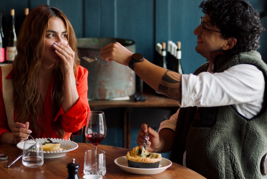 A couple eating pies on plates, the woman covering her face and the male reaching out his hand.