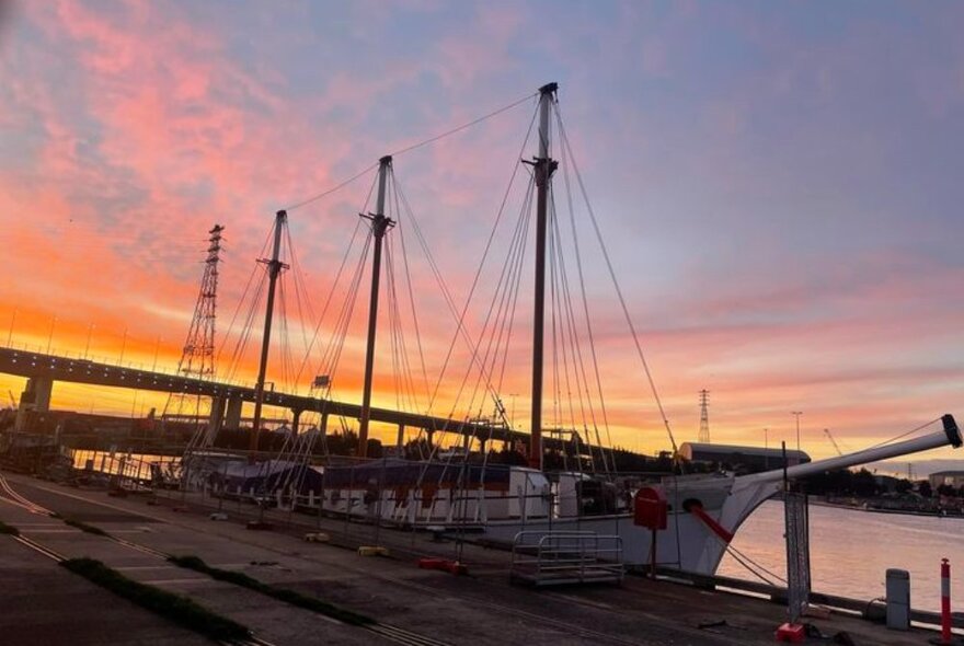 A tall ship moored at sunset with a bridge in the background.