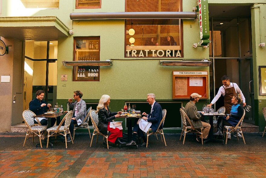 People dining at outdoor tables on wicker chairs outside green-painted trattoria with window and signage.