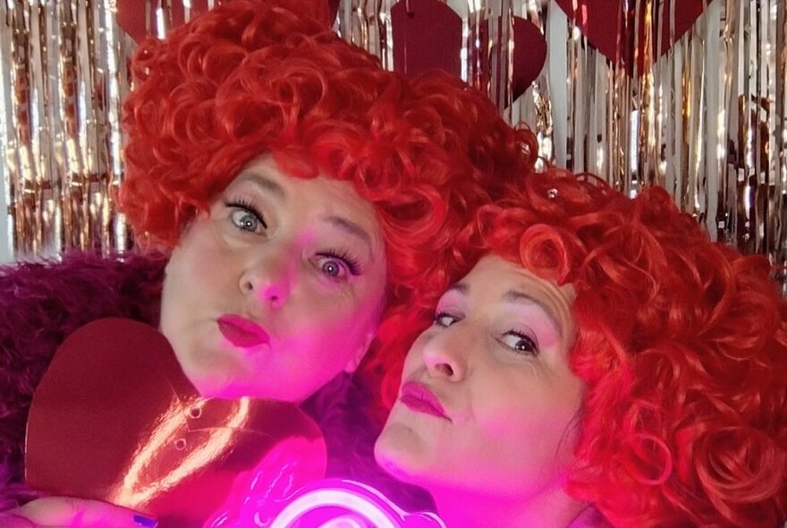 Two women wearing curly red wigs posing in front of a silver glitter curtain.