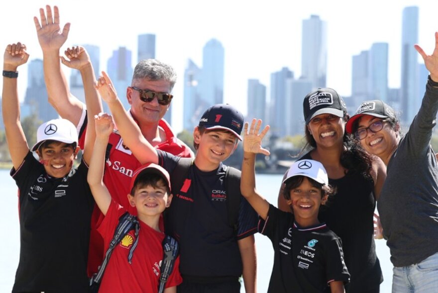 A family standing in front of Albert Park Lake waving, wearing branded caps, city skyline in the background.