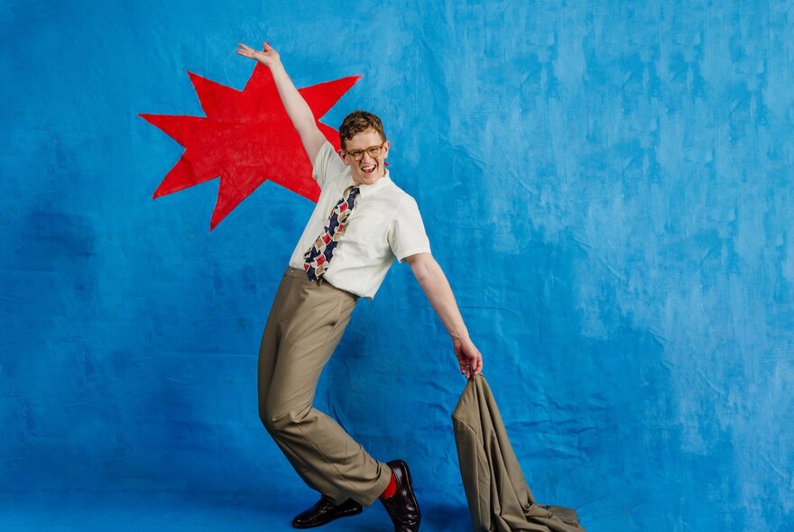 Comedian Chris Parker, posed against a blue background with one arm raised up in the air, and one arm pointing to the ground behind his back and holding his suit jacket. 