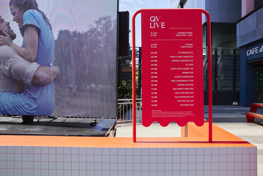 A movie showing on a large TV screen outdoors with a red program sign beside it. 