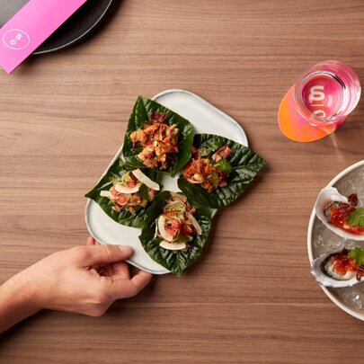 Hands taking Asian entrees from plates including oysters and betel-leaf.