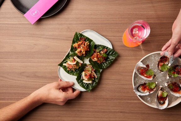 Hands taking Asian entrees from plates including oysters and betel-leaf.