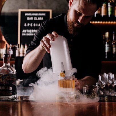 Cocktail being created and presented at a bar, with a person behind the bar and shelves of bottles visible.
