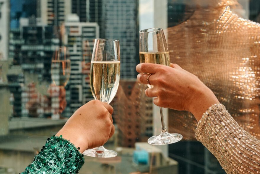 Two hands raising two flute glasses of sparkling wine against a backdrop of city skyscrapers seen though a window.