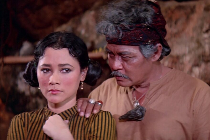 A still from an Indonesian horror film showing a young woman looking concerned, while an older man comforts her with his hand on her shoulder.
