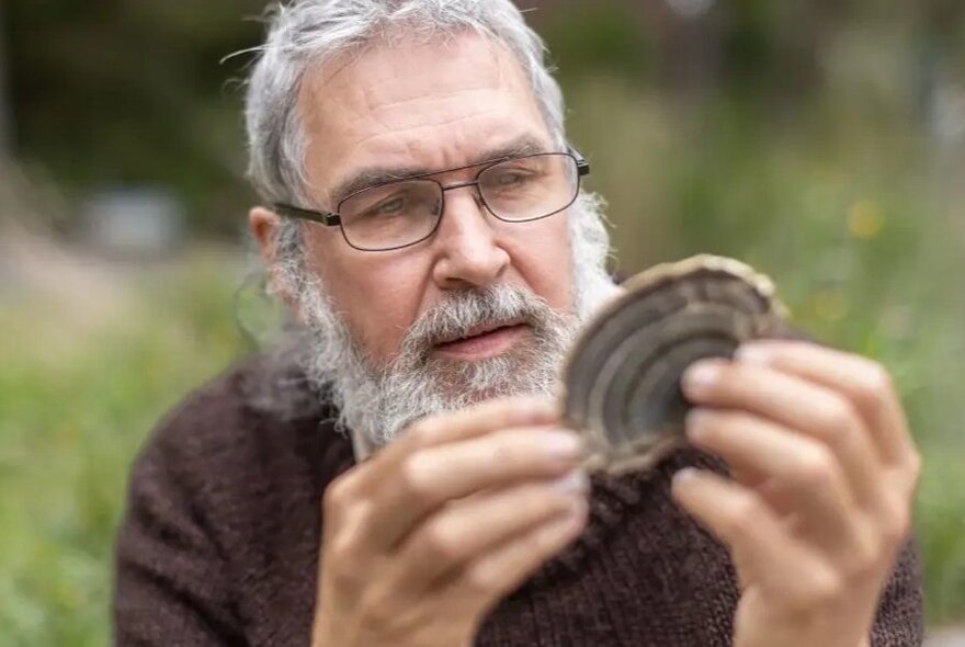 Fungi expert Dr Tom May, wearing glasses, with a greyish white beard and hair, staring intently at a mushroom he holds in his hands.