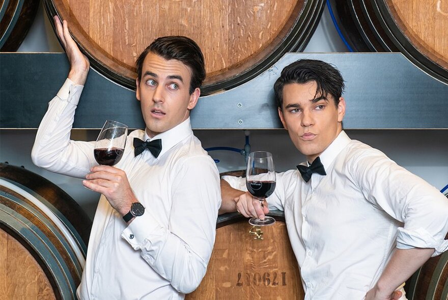 Two men dressed formally with white shirts and black bowties holding glasses of red wine and leaning on wooden barrels. 