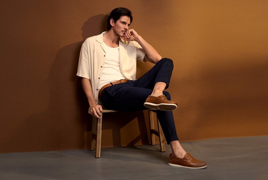A model sitting casually on a chair against a deep amber coloured wall, his legs crossed wearing tan shoes with no socks.