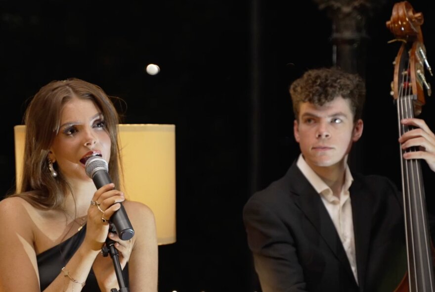 A woman singing into a microphone with a man looking on while playing the double bass. 