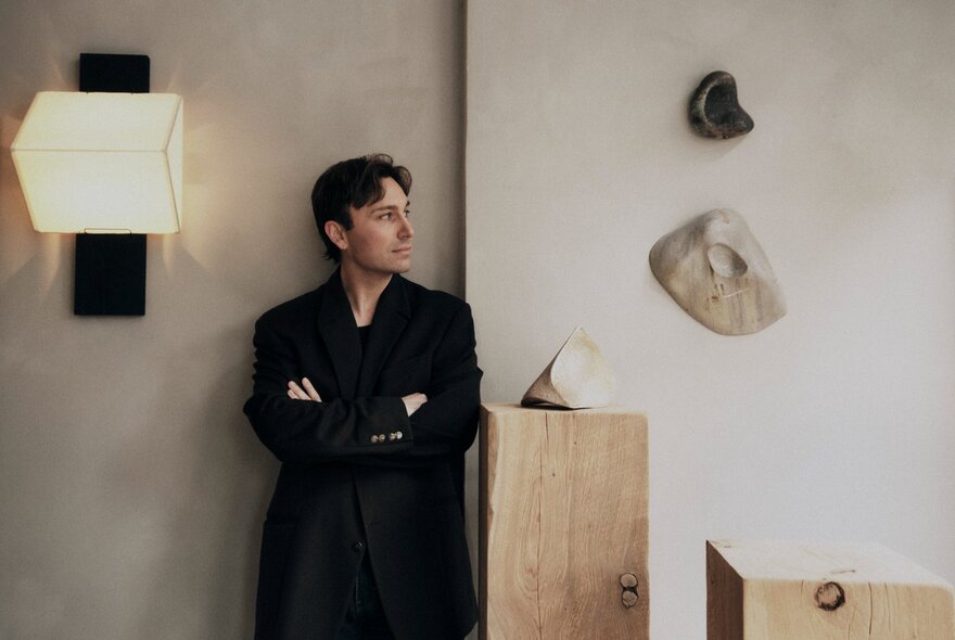 A man dressed all in black leaning against a wall with pieces of art displayed on wooden pillars next to him, or hanging on the wall.