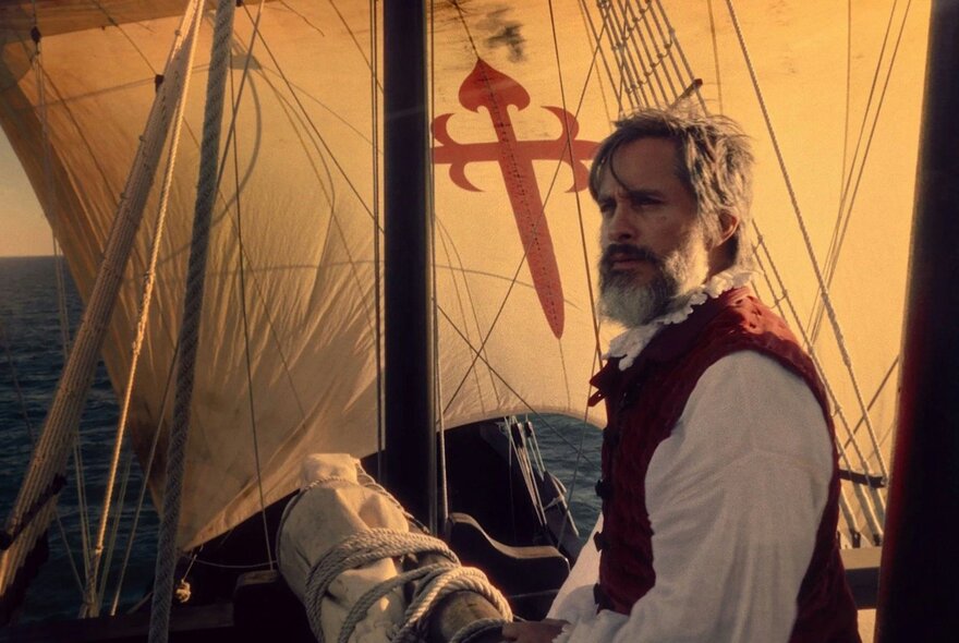 Still from a film, of a man standing on the deck of a wooden sailing ship at sunset, wearing a white shirt and a red vest. The sails and rigging of the ship fill the background.