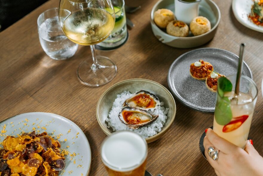 Plates of food on a restaurant dining table, with a hand holding a cocktail in a tall glass.