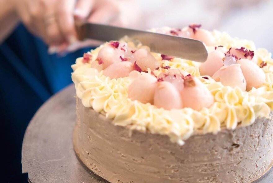 Hands holding a knife cutting a cream and pink swirl-topped cake.