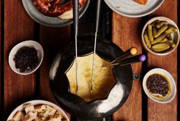 A black enamel fondue pot filled with molten cheese, fondue forks sticking out; a selection of delicious dipping food in bowls beside it.